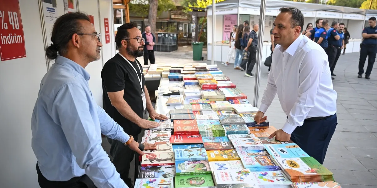 Açık havada kurulan bir kitap fuarı standında, beyaz gömlekli bir erkek, stant görevlisi olan iki kişiyle kitaplar hakkında sohbet ediyor. Uzun masanın üzerinde çok sayıda çocuk kitabı ve roman sergileniyor. Arka planda fuarı gezen insanlar ve güvenlik görevlileri görülüyor. Standın yan tarafındaki duvarda yüzde 40 indirim yazılı bir afiş asılı.