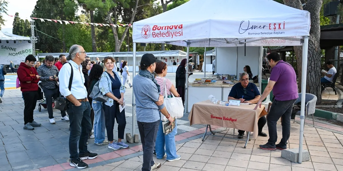 Bornova Belediyesi tarafından düzenlenen bir etkinlikte, üzerinde Bornova Belediyesi ve Bornova Belediye Başkanı Ömer Eşki'nin logolarının bulunduğu beyaz bir çadırın önünde insanlar kitap imzalatmak için sıra bekliyor. Çadırın içindeki masada oturan yazar, okurları için kitap imzalarken, mor tişörtlü bir kişi de masanın yanında duruyor. Etkinlik alanının arka planında ağaçlar ve başka stantlar görülüyor.