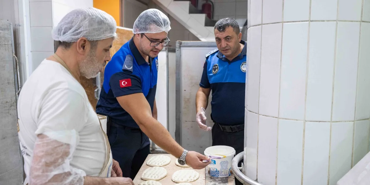 Beyaz önlük ve saç bonesi takan bir fırın çalışanı, tezgah üzerindeki hamurları hazırlarken, mavi üniformalı iki zabıta memuru denetim yapıyor. Zabıta memurlarından biri elindeki fırçayla hamurların üzerine sürülen malzemeyi kontrol ediyor.