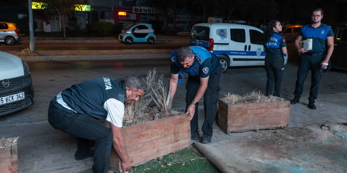Gece vakti kaldırım üzerinde çalışan Bornova Belediyesi zabıta ekipleri, ahşap saksıları yerinden kaldırıyor. Üniformalı iki zabıta memuru saksıları taşırken, arkalarında bir kadın ve bir erkek zabıta memuru onları izliyor. Arka planda üzerinde 